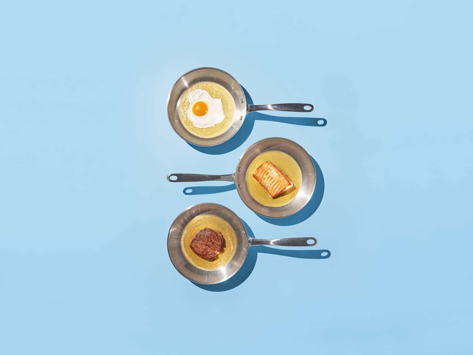 tre stekepanner på blå bunn. I stekepannene er det egg, laks og biff